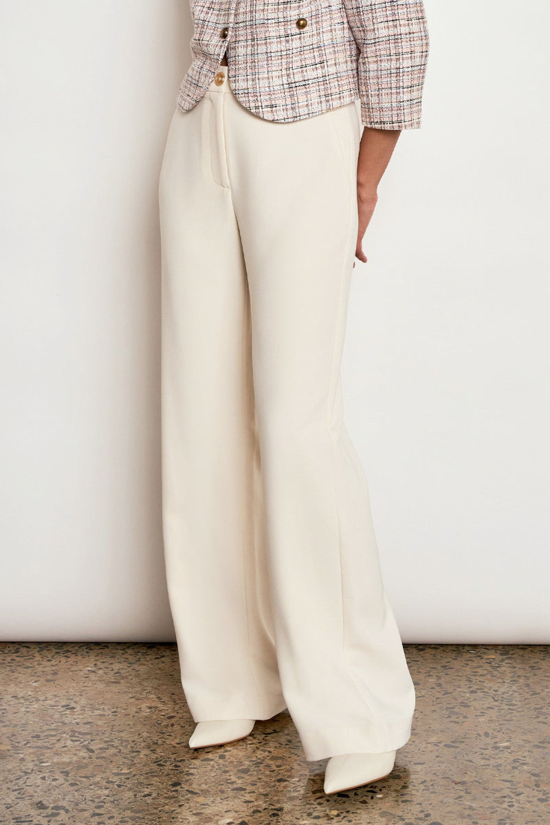 A person wearing high-waisted, wide-leg cream trousers with a plaid cropped jacket and white pointed-toe heels, standing against a plain off-white wall.