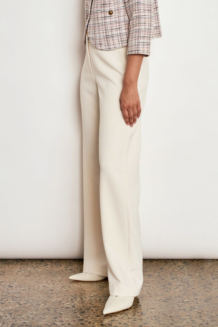 A model is shown from the waist down wearing high-waisted, wide-leg trousers in a warm cream color, paired with a plaid cropped jacket and cream pointed-toe heels, standing against a plain off-white wall.