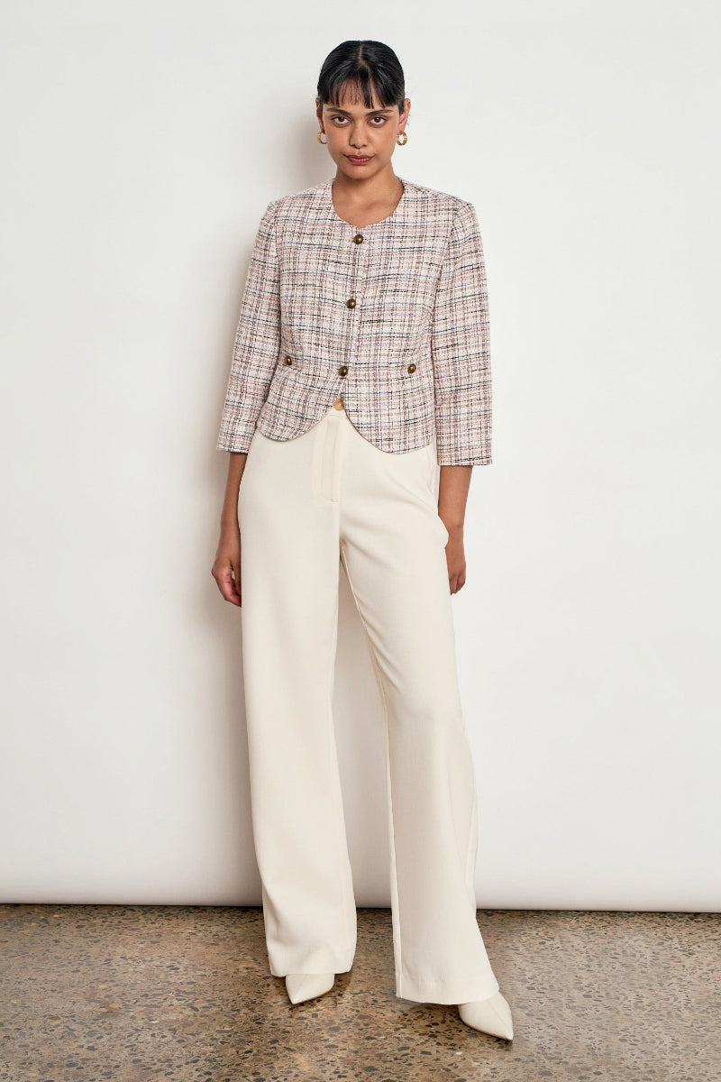 A model stands against a white wall wearing a cropped plaid jacket in beige, brown, and white with gold buttons, paired with cream wide-leg trousers.