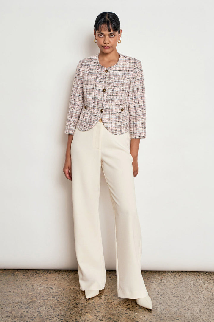 A model stands against a white wall wearing a cropped plaid jacket in beige, brown, and white with gold buttons, paired with cream wide-leg trousers.