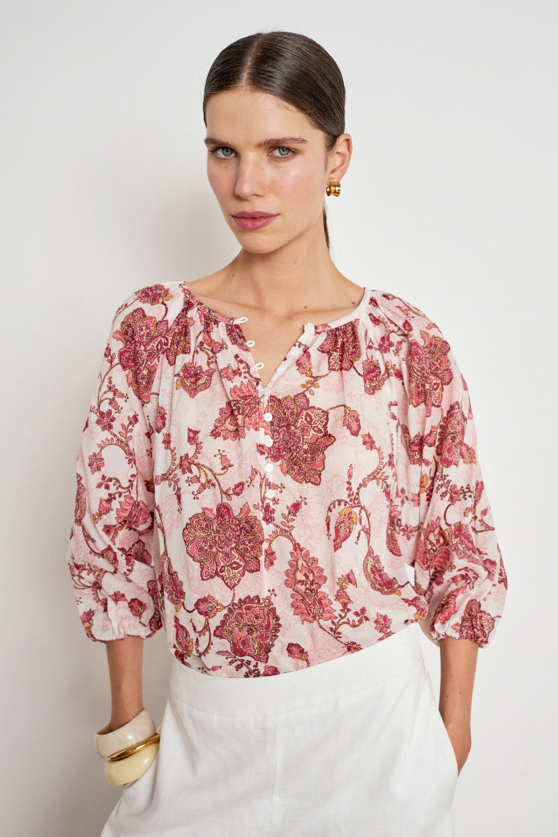 A woman wearing a light pink blouse with a red and dusty rose floral print, paired with white trousers, photographed against a neutral light gray studio background.