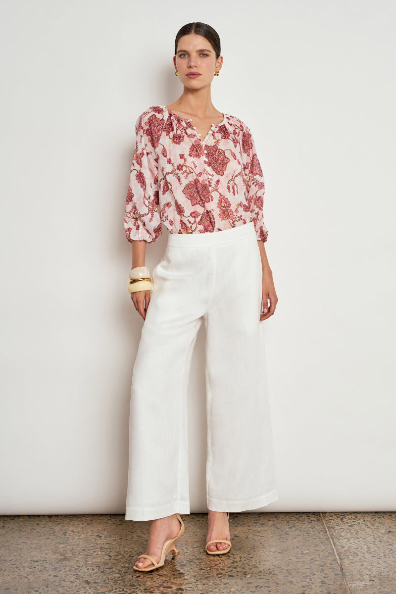 A woman stands against a plain off-white wall wearing a pink and white floral patterned blouse with puff sleeves, white wide-leg cropped pants, and beige heeled sandals.