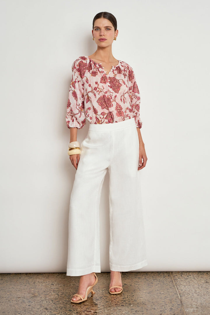 A woman stands against a plain off-white wall wearing a pink and white floral patterned blouse with puff sleeves, white wide-leg cropped pants, and beige heeled sandals.