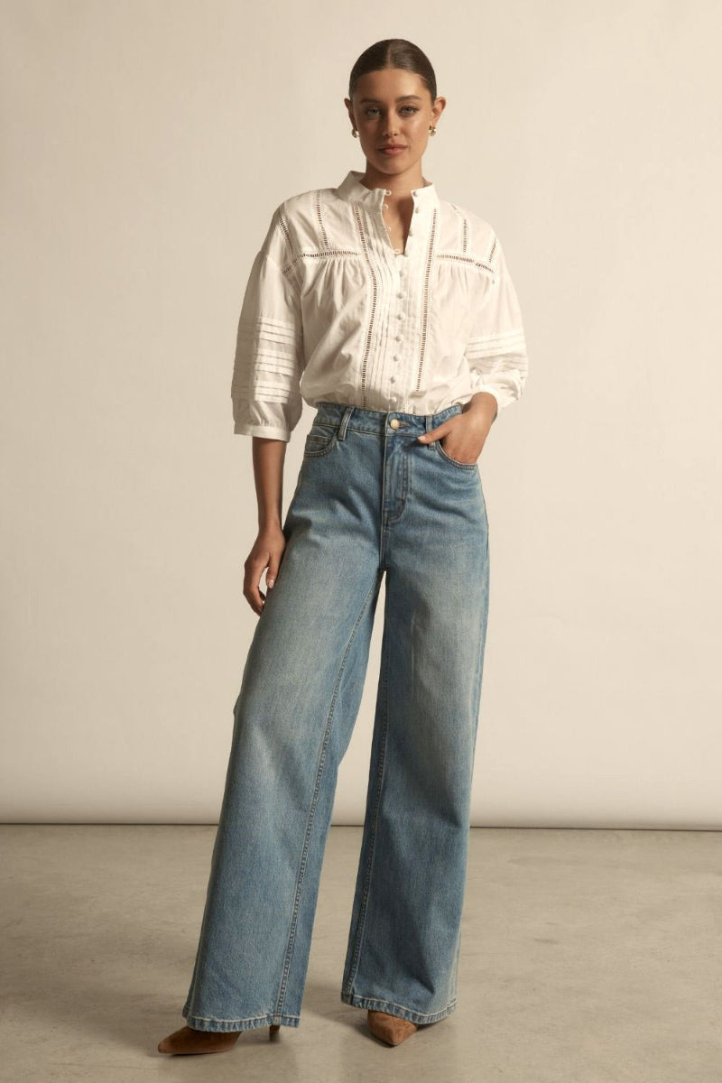 White textured blouse with short puff sleeves, delicate lace trim panels, and a row of buttons down front, slightly gathered fit with light blue baggy jeans.